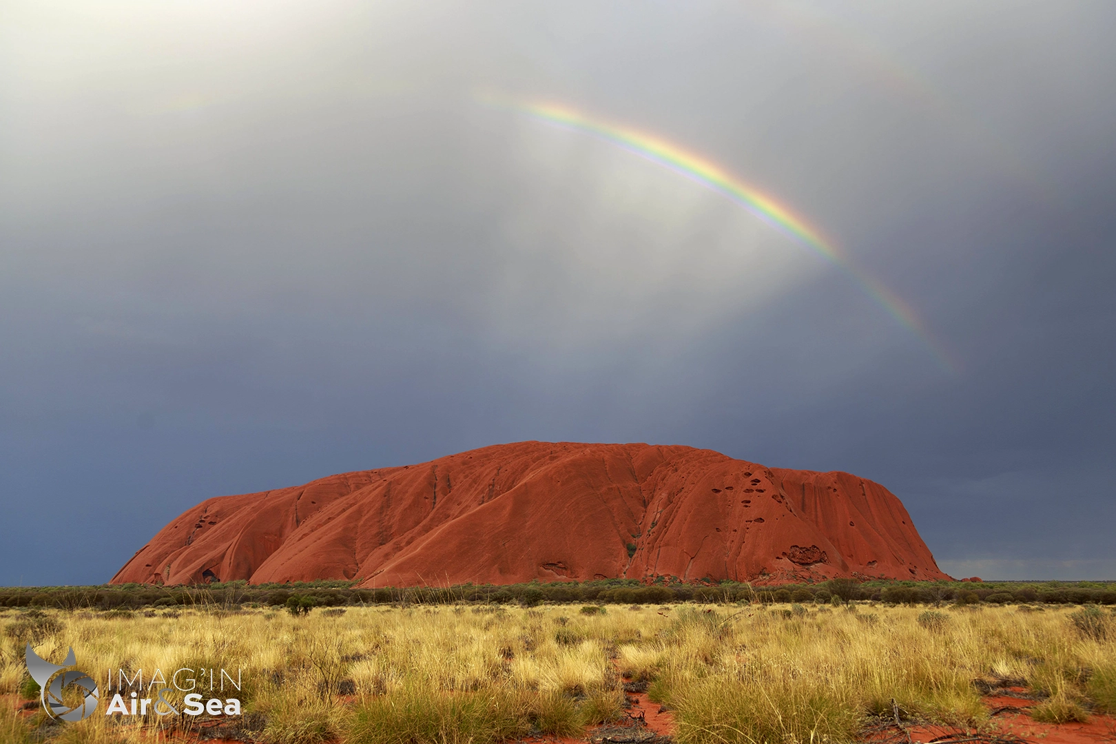 Photographie d'Ayers Rock - Uluru le rocher emblématique d'Australie sous un ciel orageux laissant apparaitre un arc en ciel