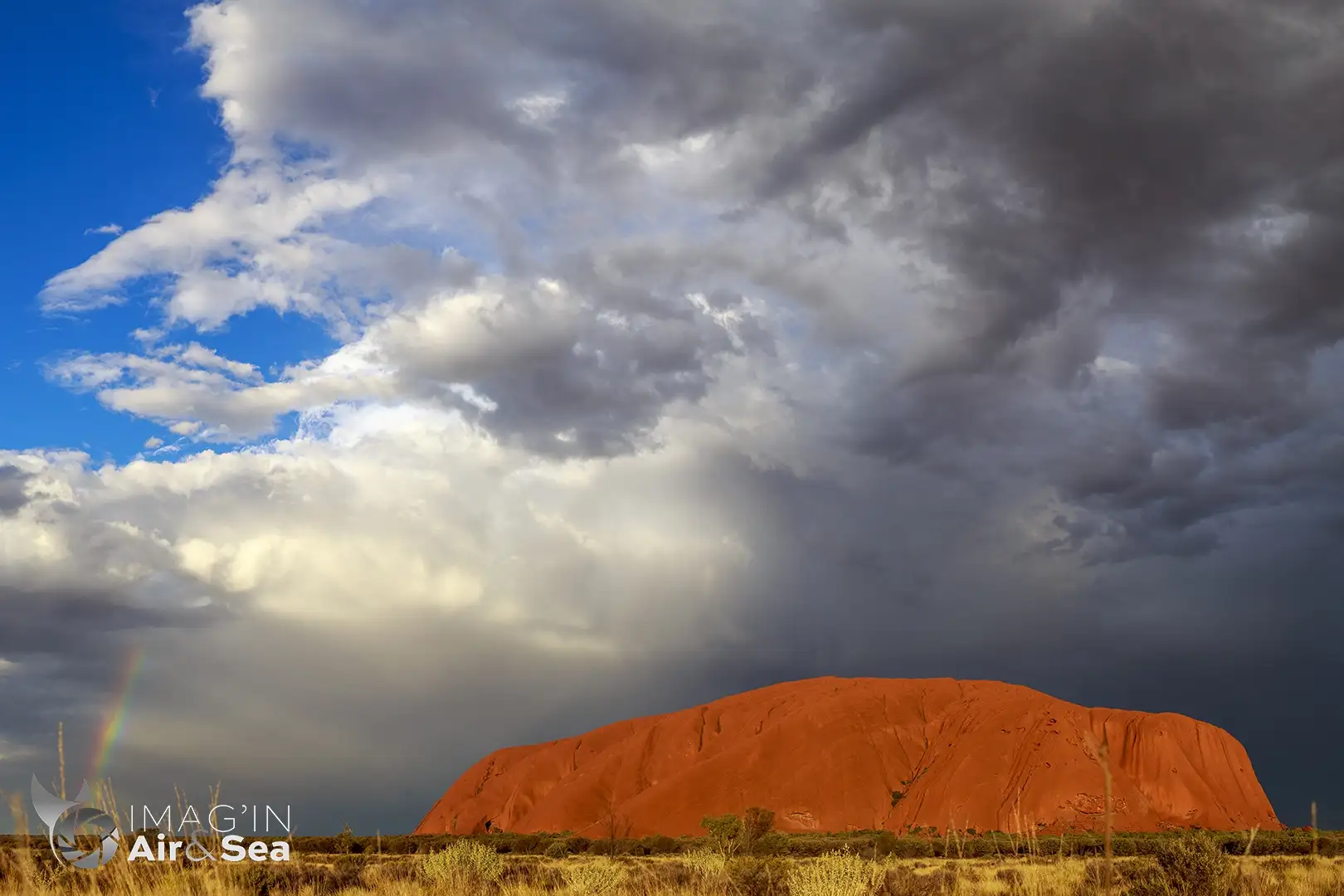 Uluru dans le centre rouge sous un ciel menaçant avec un début d'arc en ciel