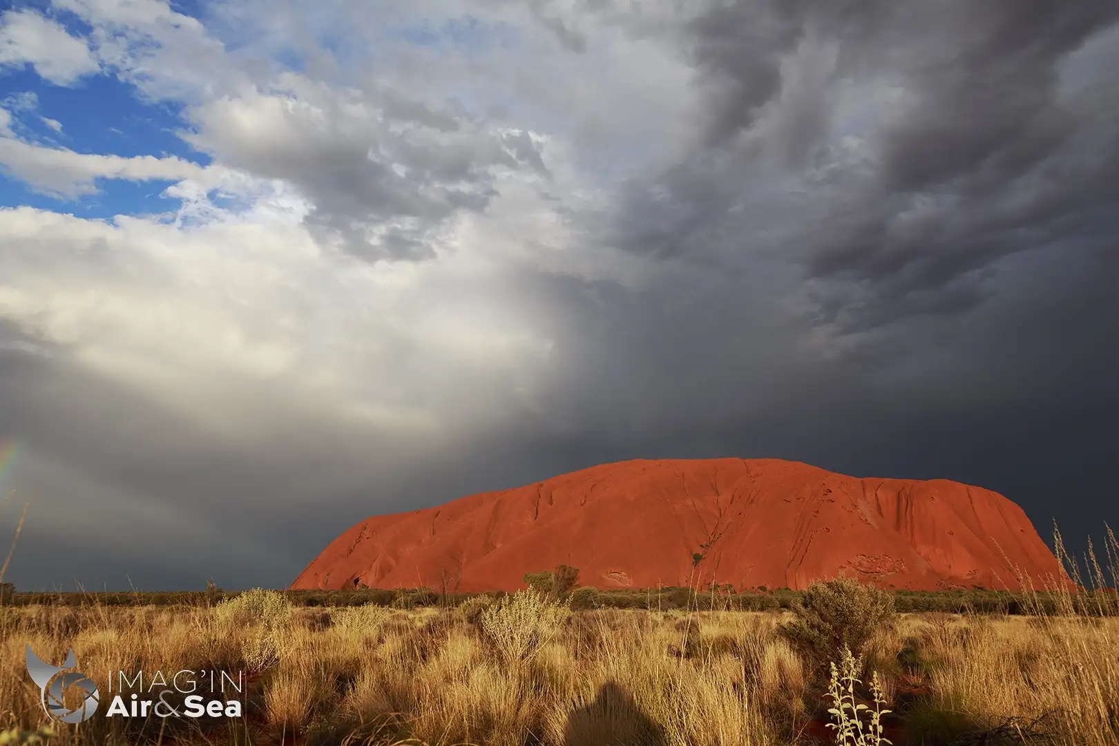 Uluru dans le centre rouge sous un ciel menaçant avec un début d'arc en ciel